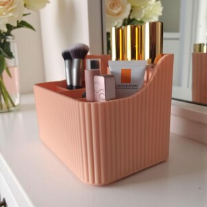 May include: A peach-coloured plastic desk organiser with a vertical ribbed texture and multiple compartments. It holds various cosmetic items, including makeup brushes, lipstick tubes, and a small white tube labelled La Roche-Posay.