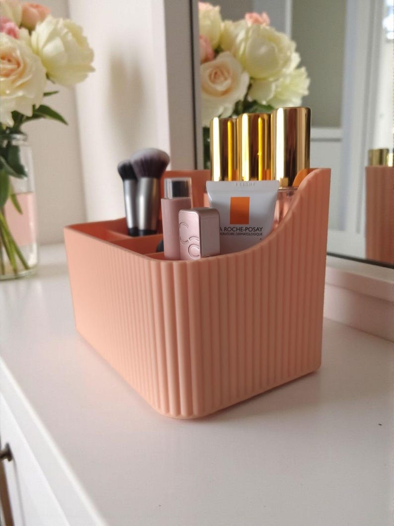 May include: A peach-coloured plastic desk organiser with a vertical ribbed texture and multiple compartments. It holds various cosmetic items, including makeup brushes, lipstick tubes, and a small white tube labelled La Roche-Posay.