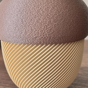 Op de afbeelding: Een decoratieve houder in de vorm van een eikel, voorzien van een getextureerde bruine dop en een crèmekleurige basis met een opvallend diagonaal ribbelpatroon. Het object heeft een glad, afgerond silhouet en een steeltje aan de bovenkant.