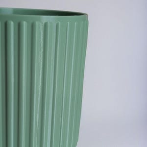 May include: A sage green cylindrical planter featuring a series of vertical, evenly spaced ridges along its exterior surface. The container has a smooth, matte finish and a wide, open top, showcasing a consistent color and a structured, ribbed design.