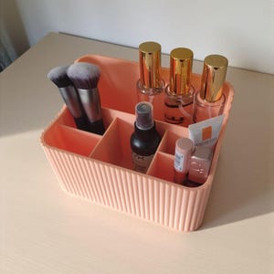 May include: A peach-coloured plastic makeup organiser with vertical ribbed detailing. The container features multiple compartments holding two makeup brushes, three clear bottles with gold caps, a black spray bottle, and two small cosmetic tubes.