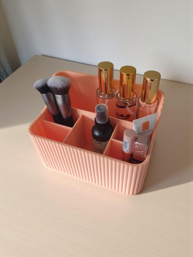 May include: A peach-coloured plastic makeup organiser with vertical ribbed detailing. The container features multiple compartments holding two makeup brushes, three clear bottles with gold caps, a black spray bottle, and two small cosmetic tubes.