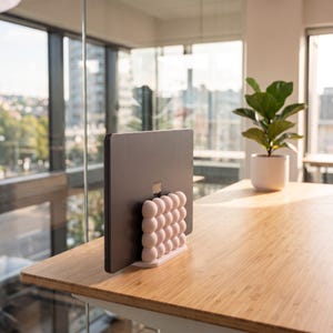May include: A dark gray laptop held upright in a light pink vertical stand. The stand features a grid of rounded, bubble-like spheres on its exterior surface, providing a stable base for the device while it is positioned in a closed, vertical orientation.