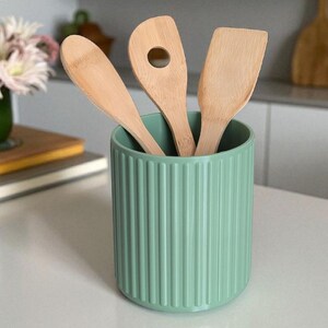 May include: A sage green cylindrical utensil holder with a vertical ribbed texture containing three light-colored wooden kitchen utensils, including a solid spoon, a spoon with a circular hole, and a flat-edged spatula.