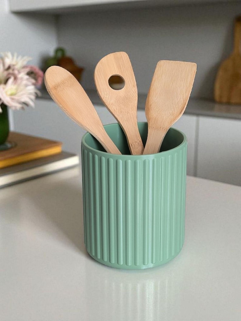 May include: A sage green cylindrical utensil holder with a vertical ribbed texture containing three light-colored wooden kitchen utensils, including a solid spoon, a spoon with a circular hole, and a flat-edged spatula.