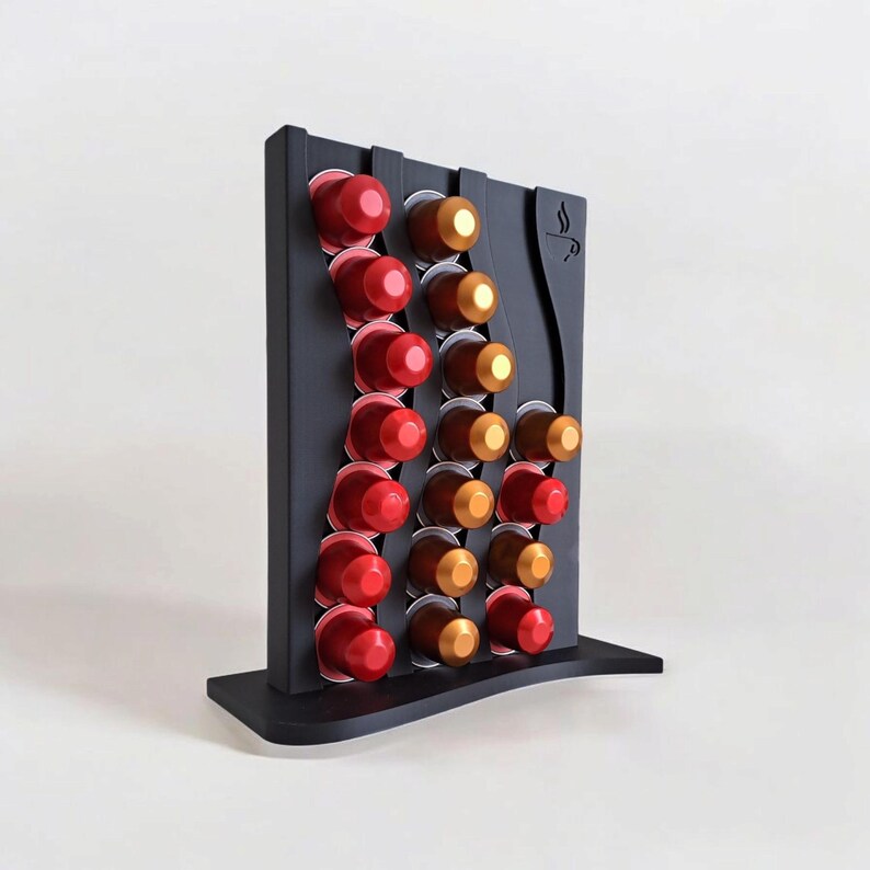 May include: A black vertical coffee pod holder with a wavy design, featuring three columns of slots filled with red and gold coffee capsules. The stand has a flat base and a small coffee cup icon engraved on the upper right side of the frame.
