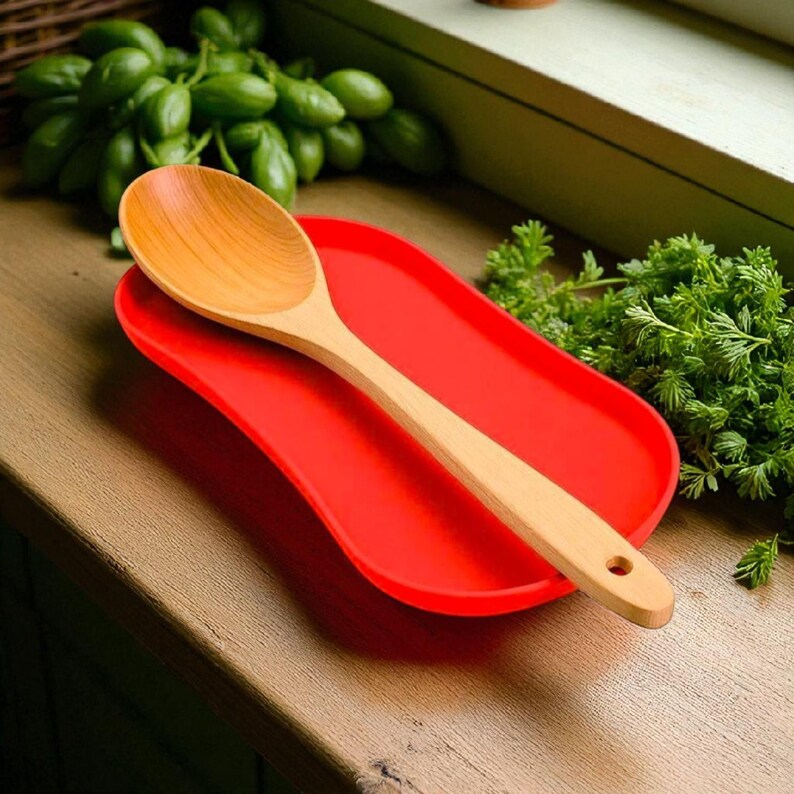 May include: A light-coloured wooden spoon with a rounded bowl and a long handle featuring a small hole at the end, resting diagonally across a bright red, oval-shaped silicone spoon rest with a smooth, matte surface finish.