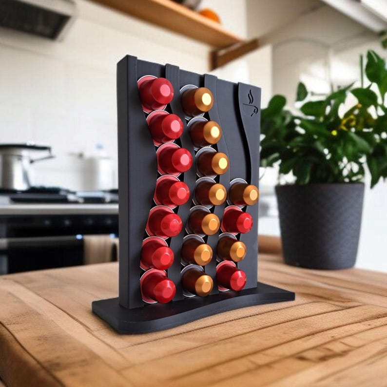 May include: A black vertical coffee pod holder with a wavy design, featuring three columns of slots filled with red and gold coffee capsules. The stand has a flat base and a small coffee cup icon engraved on the upper right side of the frame.