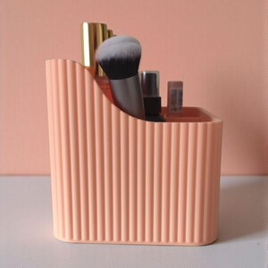 May include: A peach-coloured plastic desk organiser with a vertical ribbed texture and a curved top edge. The container holds several makeup brushes and small cosmetic bottles with metallic gold and silver caps, standing upright inside the compartments.