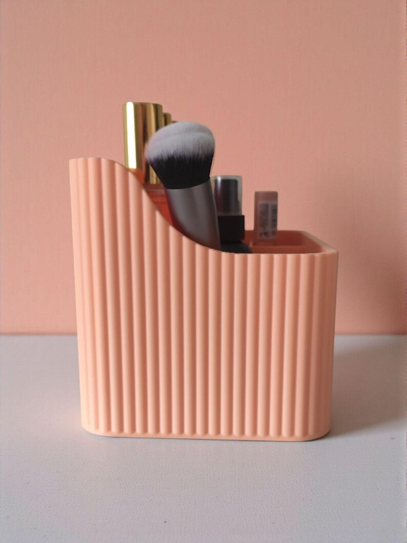 May include: A peach-coloured plastic desk organiser with a vertical ribbed texture and a curved top edge. The container holds several makeup brushes and small cosmetic bottles with metallic gold and silver caps, standing upright inside the compartments.