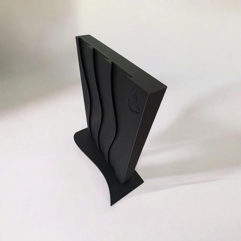 May include: A matte black vertical knife block with a wide, curved base and a tapered, upright body. The front surface features two vertical, wavy recessed channels and a small, engraved icon of a steaming cup on the upper right side.