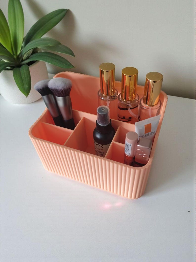 May include: A peach-coloured rectangular plastic organiser with vertical ribbed sides and multiple interior compartments. It contains two makeup brushes, three clear bottles with gold caps, one black spray bottle, and two small cosmetic tubes.
