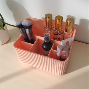 May include: A peach-coloured rectangular cosmetic organiser with vertical ribbed detailing. The container features multiple interior compartments holding two makeup brushes, three glass bottles with gold caps, a black spray bottle, and two tubes.