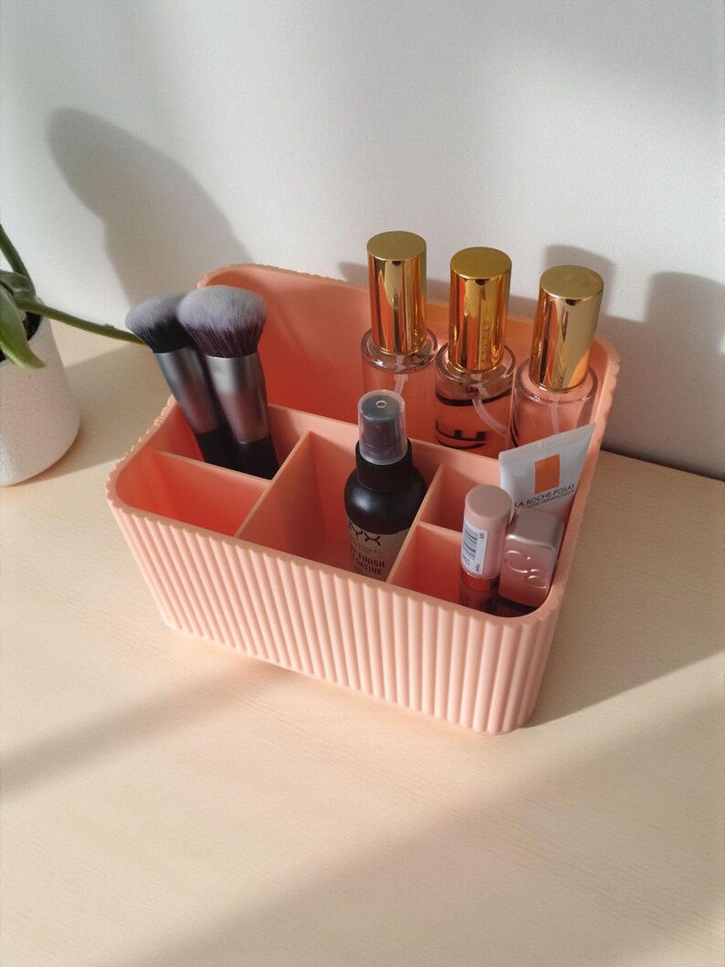 May include: A peach-coloured rectangular cosmetic organiser with vertical ribbed detailing. The container features multiple interior compartments holding two makeup brushes, three glass bottles with gold caps, a black spray bottle, and two tubes.