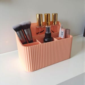 May include: A peach-coloured plastic desk organiser with a vertical ribbed texture. The rectangular container features multiple compartments holding two makeup brushes, three bottles with gold caps, a black spray bottle, and two small cosmetic tubes.