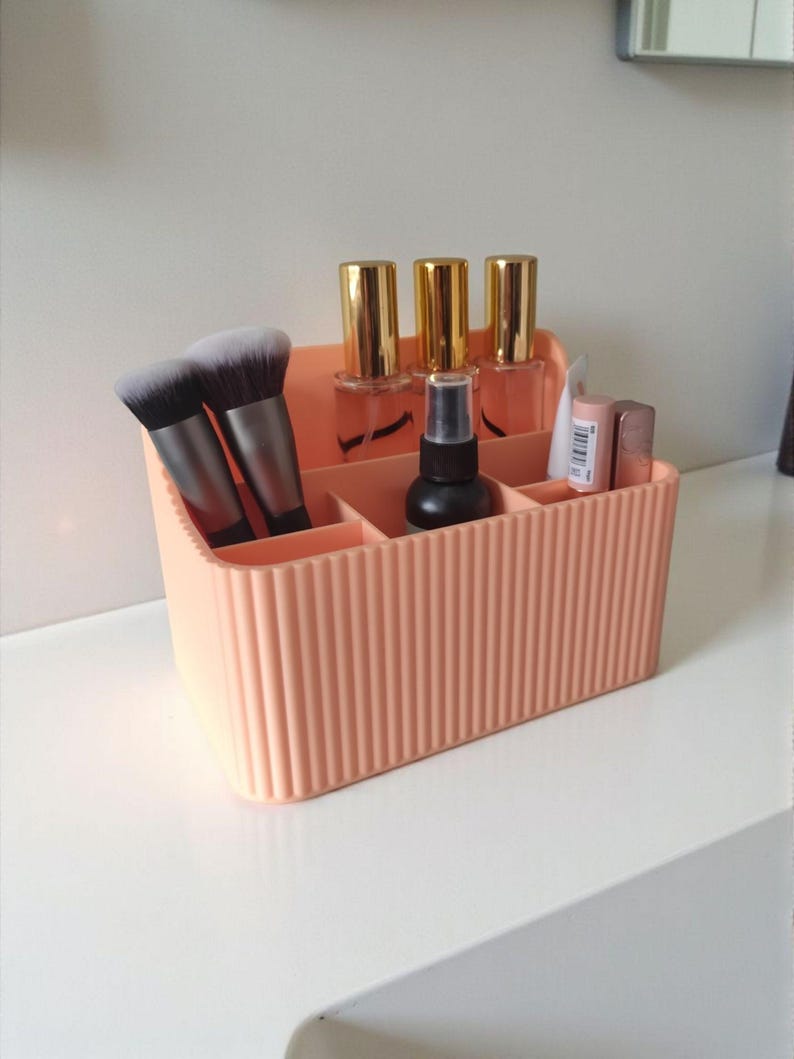 May include: A peach-coloured plastic desk organiser with a vertical ribbed texture. The rectangular container features multiple compartments holding two makeup brushes, three bottles with gold caps, a black spray bottle, and two small cosmetic tubes.