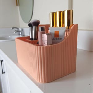 May include: A peach-coloured plastic desk organiser with a vertical ribbed texture and multiple compartments. It contains various cosmetic items, including makeup brushes, a La Roche-Posay tube, and several small bottles with gold-coloured caps.