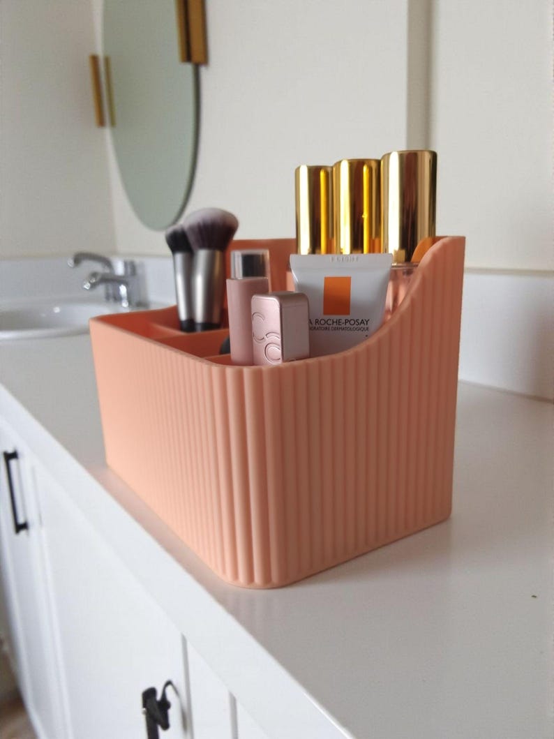 May include: A peach-coloured plastic desk organiser with a vertical ribbed texture and multiple compartments. It contains various cosmetic items, including makeup brushes, a La Roche-Posay tube, and several small bottles with gold-coloured caps.
