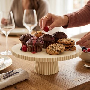 May include: A cream-colored ceramic cake stand with a fluted, ribbed design on the pedestal and the rim. The stand holds several dark chocolate muffins topped with fresh red raspberries and a stack of round, golden-brown chocolate chip cookies.