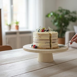 May include: A round, cream-colored cake stand with a vertically ribbed pedestal and rim, supporting a multi-layered cake frosted with white icing and topped with fresh blueberries, raspberries, and small white flowers.