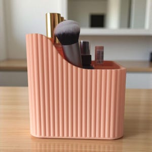 May include: A peach-coloured desk organiser with a vertical ribbed texture and a sloped top edge. The container holds several items, including a makeup brush with black and white bristles, a gold-capped bottle, and small cosmetic tubes.