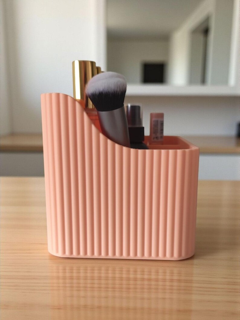 May include: A peach-coloured desk organiser with a vertical ribbed texture and a sloped top edge. The container holds several items, including a makeup brush with black and white bristles, a gold-capped bottle, and small cosmetic tubes.