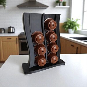 Puede incluir: Un soporte vertical negro para c&aacute;psulas de caf&eacute; con dise&ntilde;o ondulado, que cuenta con dos columnas para un total de ocho c&aacute;psulas de caf&eacute; marrones. El soporte tiene una base rectangular plana y un acabado mate y liso.