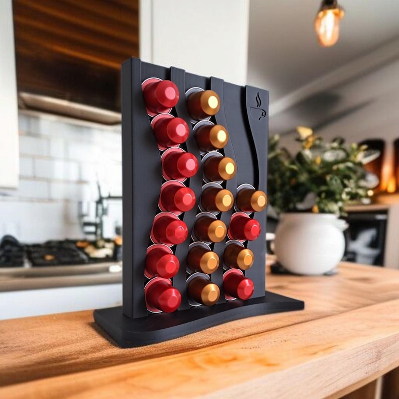 May include: A black vertical coffee pod holder with a wavy design, featuring three columns of slots filled with red and gold coffee capsules. The stand has a flat base and a small coffee cup icon engraved on the upper right side of the frame.