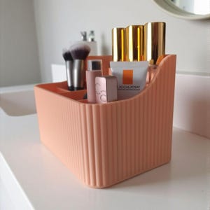 May include: A peach-coloured plastic desk organiser with a vertical ribbed texture. The container features multiple compartments holding various cosmetic items, including makeup brushes, lip products, and a tube labelled La Roche-Posay.