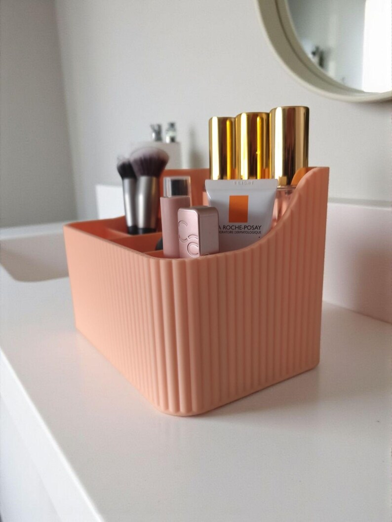 May include: A peach-coloured plastic desk organiser with a vertical ribbed texture. The container features multiple compartments holding various cosmetic items, including makeup brushes, lip products, and a tube labelled La Roche-Posay.