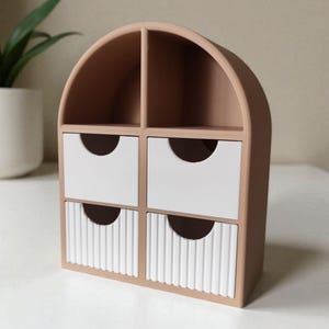 May include: A desktop organizer with a tan arched frame featuring two open top compartments and four drawers. The top two drawers are smooth white, while the bottom two drawers feature a vertical ribbed texture, each with a semi-circular cutout.