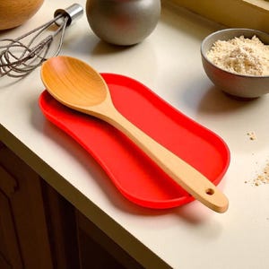 May include: A bright red silicone spoon rest with an elongated oval shape and a smooth surface. A light-coloured wooden spoon with a rounded bowl and a small hole at the end of the handle rests diagonally across the centre of the red spoon rest.