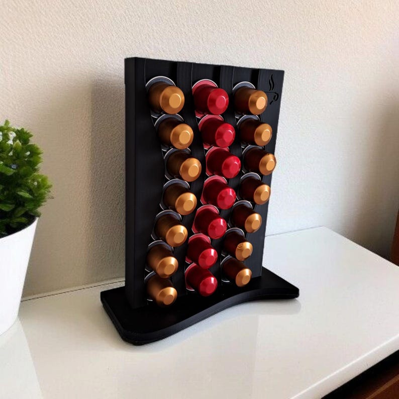 May include: A black vertical coffee pod holder with a wavy design, featuring three columns of slots filled with gold, brown, and red coffee capsules. The top right corner displays a small engraved icon of a coffee cup with steam rising from it.