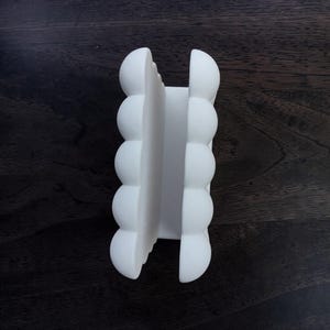 May include: A matte white napkin holder featuring a vertical design with two parallel sides. Each side is composed of five stacked, rounded, semi-spherical shapes that create a textured, scalloped edge along the length of the object.
