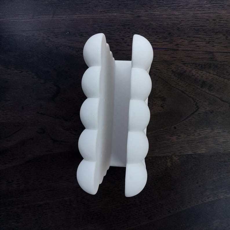 May include: A matte white napkin holder featuring a vertical design with two parallel sides. Each side is composed of five stacked, rounded, semi-spherical shapes that create a textured, scalloped edge along the length of the object.