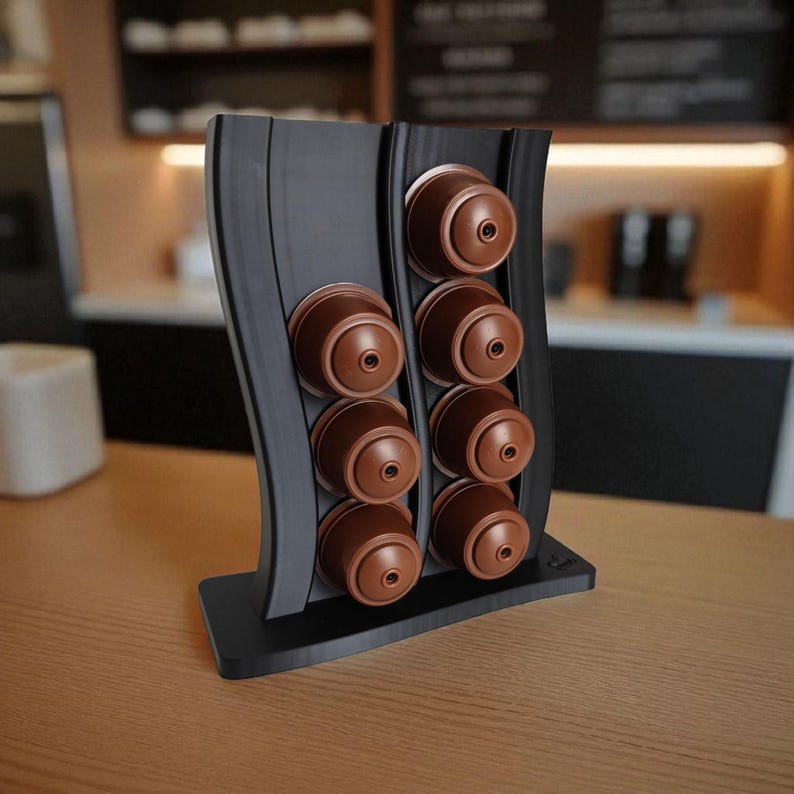 Puede incluir: Un soporte vertical negro para c&aacute;psulas de caf&eacute; con dise&ntilde;o ondulado, que cuenta con dos columnas para un total de ocho c&aacute;psulas de caf&eacute; marrones. El soporte tiene una base rectangular plana y un acabado mate liso para el almacenamiento.