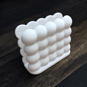 May include: A white napkin holder constructed from a grid of spherical shapes. The structure features a repeating pattern of smooth, matte white spheres arranged in a rectangular formation, creating a three-dimensional bubble-like appearance.