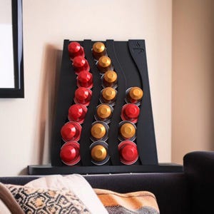 May include: A black coffee pod holder with a wavy design, featuring three vertical columns holding red and gold coffee capsules. The top right corner of the holder displays a small engraved icon of a coffee cup with steam rising from it.