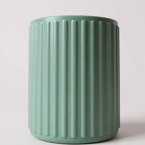 May include: A sage green cylindrical cup featuring a vertical ribbed texture along the exterior surface. The vessel has a smooth, matte finish and a consistent color throughout, with a simple open top and a flat base, presenting a clean design.