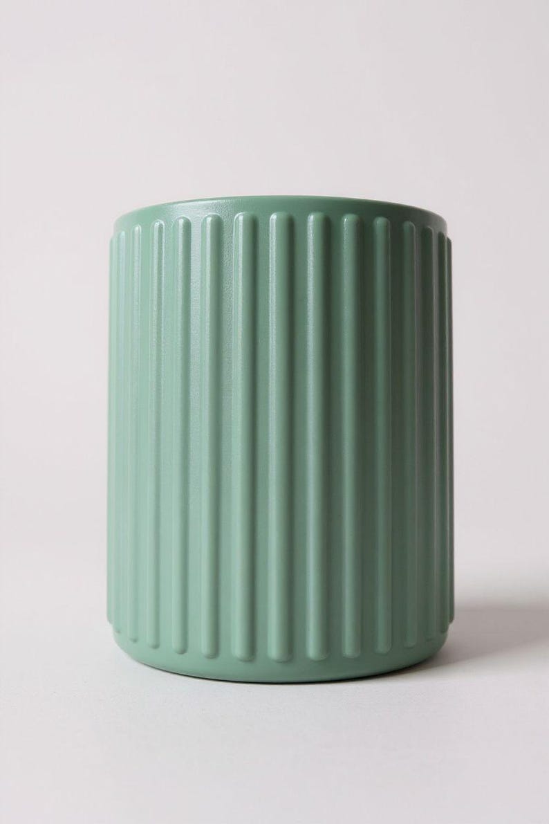 May include: A sage green cylindrical cup featuring a vertical ribbed texture along the exterior surface. The vessel has a smooth, matte finish and a consistent color throughout, with a simple open top and a flat base, presenting a clean design.
