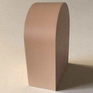 May include: A solid, light beige, arch-shaped object with a smooth, matte finish. The item features a flat base and a curved top, standing upright with a uniform, opaque color throughout its structure, presenting a clean, geometric form.