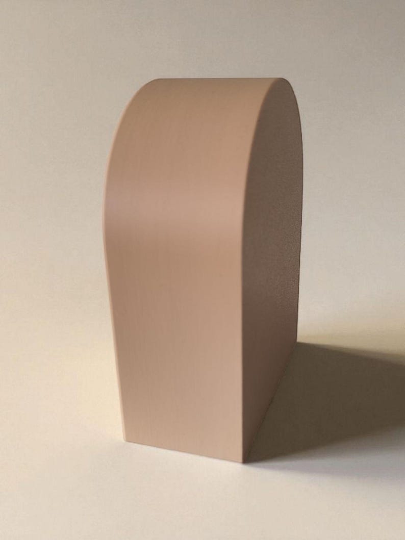 May include: A solid, light beige, arch-shaped object with a smooth, matte finish. The item features a flat base and a curved top, standing upright with a uniform, opaque color throughout its structure, presenting a clean, geometric form.
