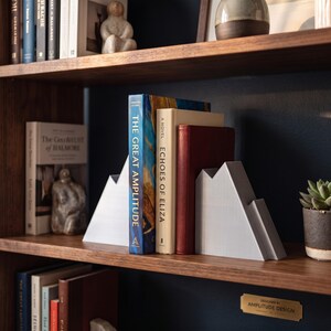 Peut inclure: Deux serre-livres gris clair en forme de montagne fabriqu&eacute;s dans un mat&eacute;riau lisse, plac&eacute;s sur une &eacute;tag&egrave;re en bois pour soutenir des livres dont les titres The Great Amplitude et Echoes of Eliza, devant un mur bleu fonc&eacute; en arri&egrave;re-plan.