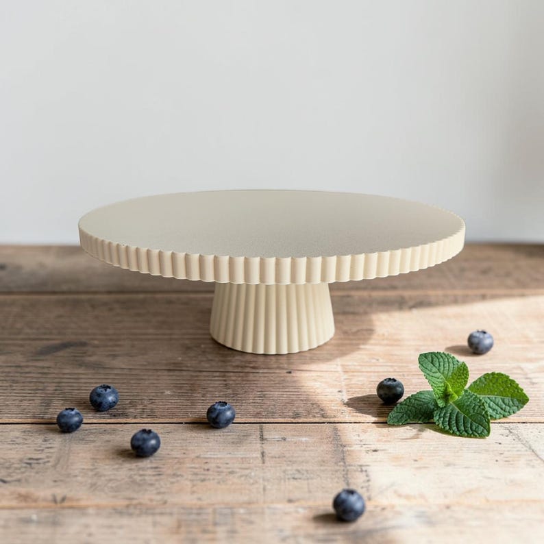 May include: A cream-colored cake stand with a circular top and a pedestal base, both featuring a vertical fluted texture. The stand is positioned on a wooden surface alongside several scattered fresh blueberries and a sprig of green mint leaves.