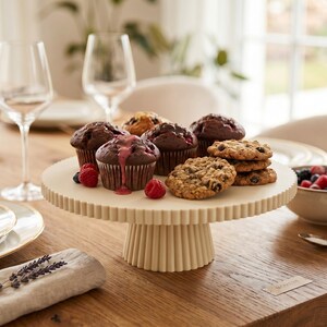 May include: A cream-colored, fluted pedestal cake stand holding several chocolate muffins, one with pink icing, and a stack of chocolate chip cookies. Fresh raspberries are scattered on the surface of the stand next to the baked goods.