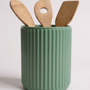 May include: A sage green cylindrical utensil holder with a vertical ribbed texture. Three light brown wooden kitchen utensils, including a spoon, a slotted spoon, and a flat spatula, are placed inside the container, extending upwards.