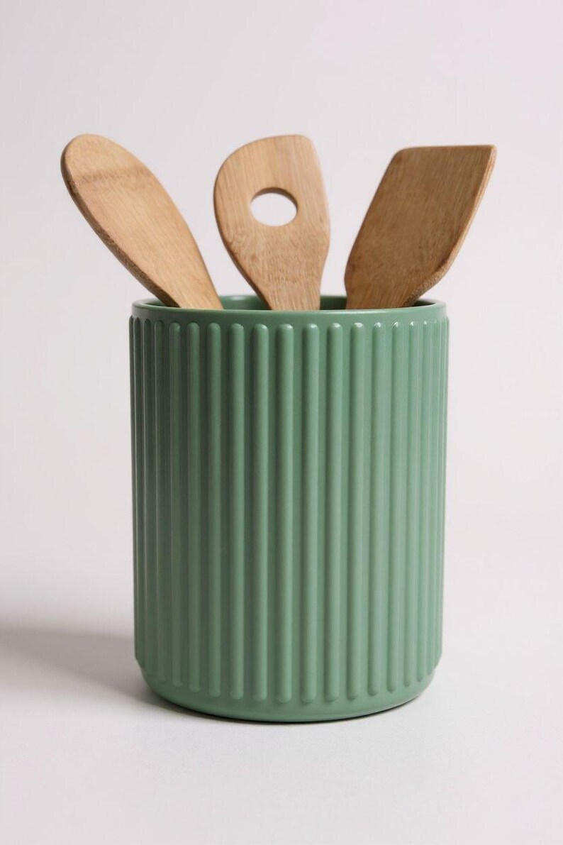May include: A sage green cylindrical utensil holder with a vertical ribbed texture. Three light brown wooden kitchen utensils, including a spoon, a slotted spoon, and a flat spatula, are placed inside the container, extending upwards.