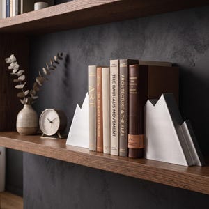 Peut inclure: Une paire de serre-livres en m&eacute;tal argent&eacute; en forme de sommets montagneux, plac&eacute;s sur une &eacute;tag&egrave;re en bois pour soutenir une collection de livres reli&eacute;s, dont les titres incluent The Bauhaus Movement et Architecture and the Alps.