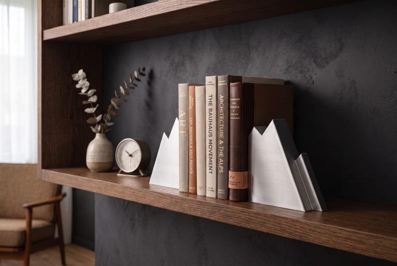 Peut inclure: Une paire de serre-livres en m&eacute;tal argent&eacute; en forme de sommets montagneux, plac&eacute;s sur une &eacute;tag&egrave;re en bois pour soutenir une collection de livres reli&eacute;s, dont les titres incluent The Bauhaus Movement et Architecture and the Alps.