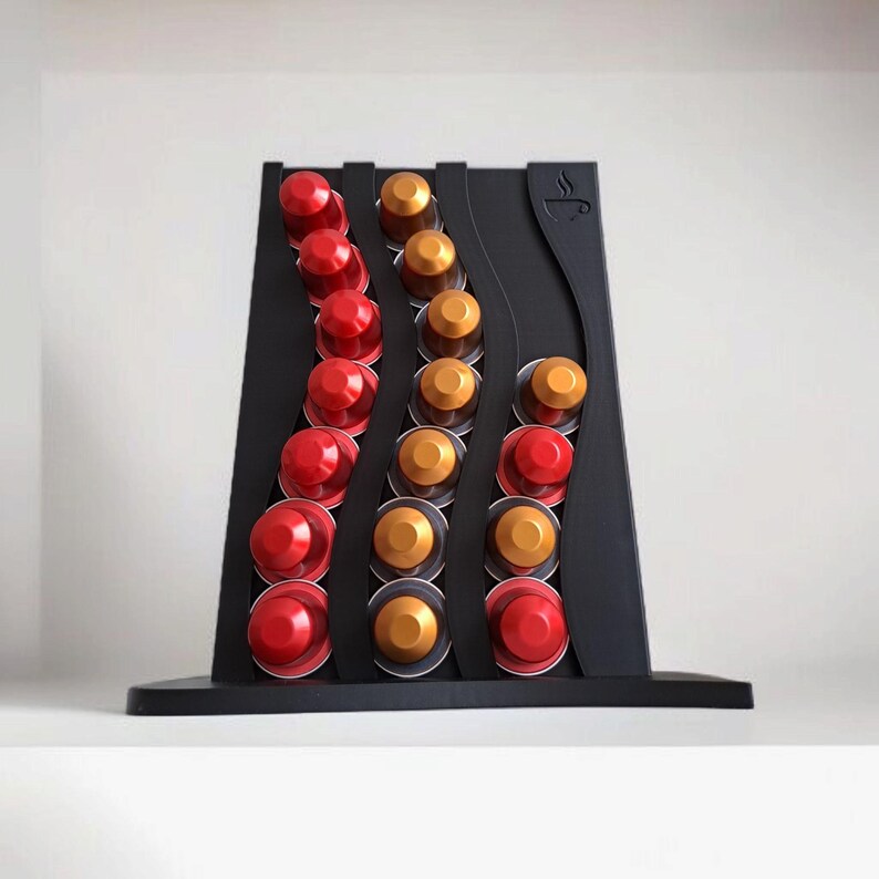 May include: A black coffee pod holder with a wavy design featuring three vertical columns. The holder contains red and gold coffee capsules arranged in the slots. A small icon of a coffee cup with steam is embossed on the top right corner of the frame.